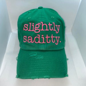 May include: Green baseball cap with distressed edges and pink embroidery that reads "slightly saditty."