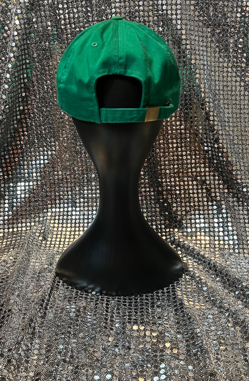 Op de afbeelding: Een groene baseballpet met een bruine band op een zwart manekin. De pet is op een zilveren, glinsterende achtergrond.