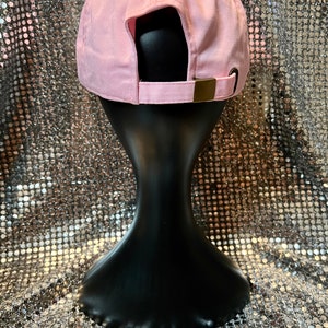 Puede incluir: Una gorra de b&eacute;isbol rosa con una correa ajustable marr&oacute;n en la parte posterior. La gorra est&aacute; en una cabeza de maniqu&iacute; negra.