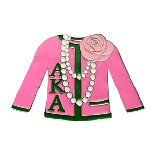 May include: A pink jacket pin with green trim and the letters AKA. A white pearl necklace and a pink rose are also featured. The pin is a decorative accessory.