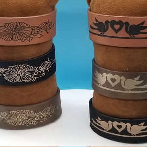 May include: Four leather bracelets with different designs. Two bracelets feature a floral design, and two bracelets feature a design of two birds with a heart in the middle. The bracelets are stacked on top of each other, with the brown leather bracelets on the bottom and the black leather bracelets on top.