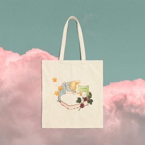 May include: A cream-colored canvas tote bag featuring a printed illustration of a light brown rabbit wearing a blue jacket, standing beside a bed with a green headboard, accompanied by three small orange stars and a small cluster of dark red flowers.