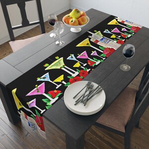 May include: Black table runner with a colorful design of martini glasses and tropical flowers. The runner is on a dark wood table with a white plate and silverware.