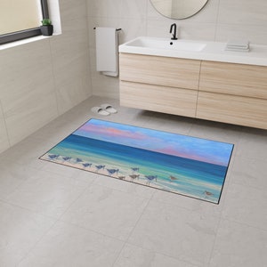 Op de afbeelding: Een rechthoekige vloermat met een bedrukte kustsc&egrave;ne. Het ontwerp toont een zandstrand met verschillende kleine strandvogels bij de waterkant, een turquoise oceaan en een lucht met zachtroze en blauwe wolken tijdens de zonsondergang.