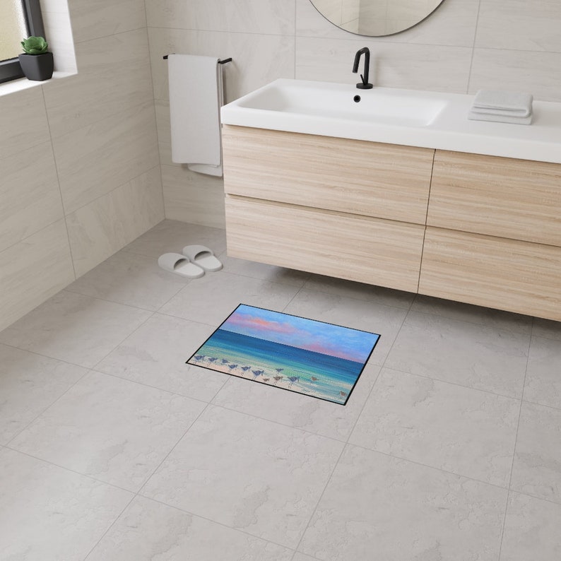 Op de afbeelding: Een rechthoekige vloermat met een geprinte afbeelding van een strandsc&egrave;ne bij zonsondergang. Het ontwerp toont een zanderige kustlijn met kleine vogels, turquoise zeewater en een lucht vol zachte roze en blauwe wolken, omlijst door een dunne zwarte rand.
