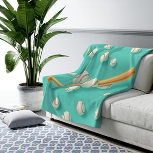 May include: A soft throw blanket in a vibrant turquoise colour featuring a repeating pattern of white baseballs with red stitching and a wooden baseball bat positioned diagonally across the fabric, draped over the corner of a white sofa.