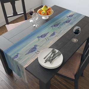 May include: A rectangular table runner with a watercolor design of shorebirds in shades of blue and gray. The runner is placed on a dark wooden table, with a bowl of fruit and glassware visible.