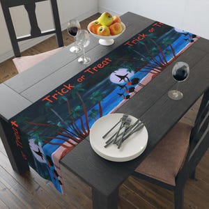 May include: A rectangular table runner featuring a nighttime beach scene with palm trees, a full moon, and silhouettes of birds. The text "Trick or Treat" is printed in orange across the runner. The runner is displayed on a dark wooden table.