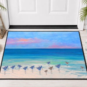 Op de afbeelding: Een rechthoekige deurmat met een kusttafereel met een groep kleine strandvogels op een lichtgekleurd zandstrand. De achtergrond toont een turquoise oceaan die samenkomt met een blauwe lucht met zachte roze en oranje zonsondergangwolken.
