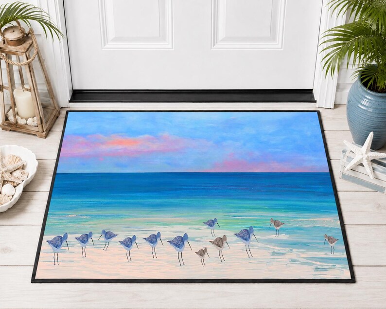 Op de afbeelding: Een rechthoekige deurmat met een kusttafereel met een groep kleine strandvogels op een lichtgekleurd zandstrand. De achtergrond toont een turquoise oceaan die samenkomt met een blauwe lucht met zachte roze en oranje zonsondergangwolken.