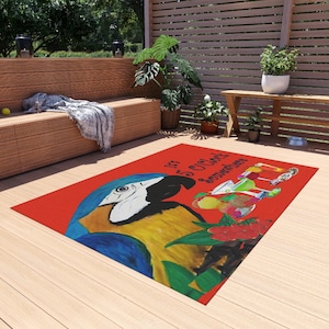 May include: A red outdoor rug featuring a vibrant illustration of a blue and yellow macaw parrot. The rug has the text "It's 5 O'Clock Somewhere" and a colorful cocktail design. The rug is placed on a wooden deck.