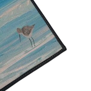 Op de afbeelding: Een rechthoekige mat met een gedrukte illustratie van een kleine bruine strandloper die op een lichtblauw en wit gestructureerd oppervlak staat dat lijkt op water of zand. De mat heeft een dikke zwarte gestikte rand langs de zichtbare zijde.