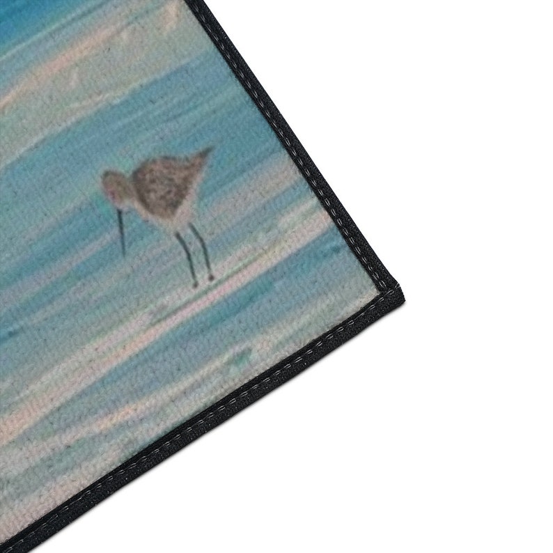 Op de afbeelding: Een rechthoekige mat met een gedrukte illustratie van een kleine bruine strandloper die op een lichtblauw en wit gestructureerd oppervlak staat dat lijkt op water of zand. De mat heeft een dikke zwarte gestikte rand langs de zichtbare zijde.