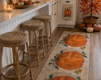 May include: A rectangular runner rug featuring three large orange pumpkins with green stems and leaves printed on a light beige background. The rug is positioned on a wooden floor in a kitchen decorated with autumn-themed decor and furniture.