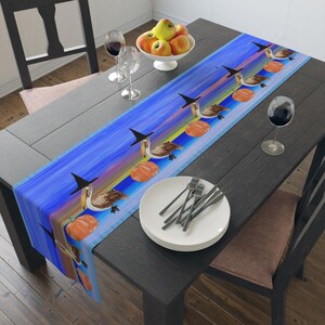 May include: A blue table runner with a whimsical Halloween design. It features pelicans wearing witch hats, orange pumpkins, and a rainbow-colored background. The runner is placed on a dark wooden table, with a white plate, silverware, and wine glasses.