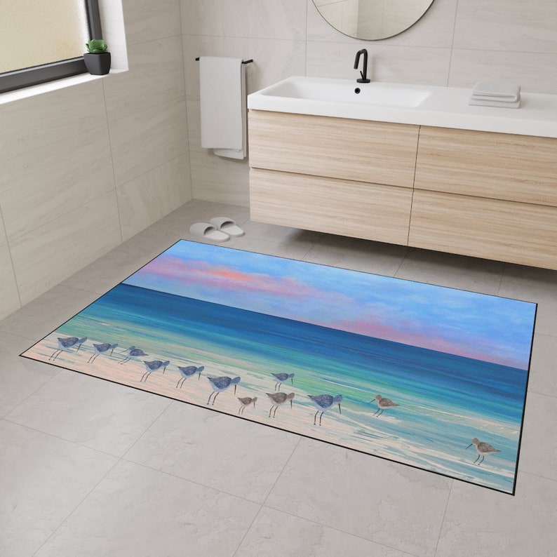 Op de afbeelding: Een rechthoekige vloermat met een bedrukte kustsc&egrave;ne van een rij kleine strandvogels die langs een zandstrand lopen. De afbeelding toont een verloop van blauw zeewater, wit schuim en een zachte roze en blauwe lucht bij zonsondergang.