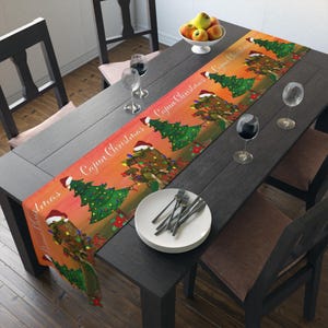 May include: A colourful table runner with a Cajun Christmas design. The runner displays a gradient of orange and red with cartoon alligators wearing Santa hats and decorated Christmas trees. The words "Cajun Christmas" are printed along the length of the runner.