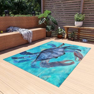 Puede incluir: Una alfombra rectangular para exteriores con una pintura de acuarela de un cangrejo azul sobre un fondo turquesa. El cangrejo tiene pinzas y cuerpo detallados, con una paleta de colores azul claro y morado. La alfombra está colocada en una terraza de madera.