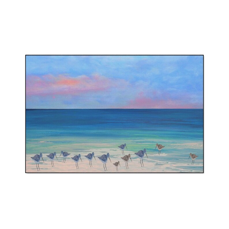 Op de afbeelding: Een rechthoekig schilderij met een groep kleine strandvogels die op een licht zandstrand aan de waterkant staan. De sc&egrave;ne toont een kalme blauwe oceaan onder een zachte lucht met roze en blauwe wolken, een sereen kustlandschap.