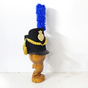 May include: A black hat with a gold emblem, gold rope trim, and a blue plume. The hat is displayed on a wooden stand with a carved design. The hat has a black brim and a gold tassel.