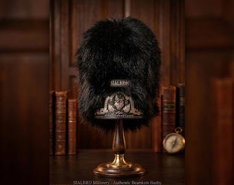 May include: A tall black bearskin busby hat featuring a prominent silver-toned metal badge at the front base. The badge displays an intricate crest design. A small rectangular label with the text SIALBRO is centered on the front of the fur.