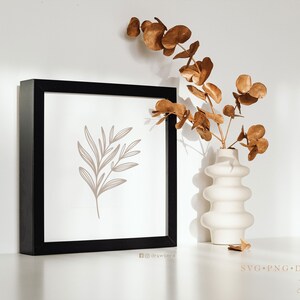 May include: A black framed print featuring a simple line drawing of a branch with leaves in a light brown colour. The print is sitting on a white surface next to a white vase with dried brown eucalyptus branches.