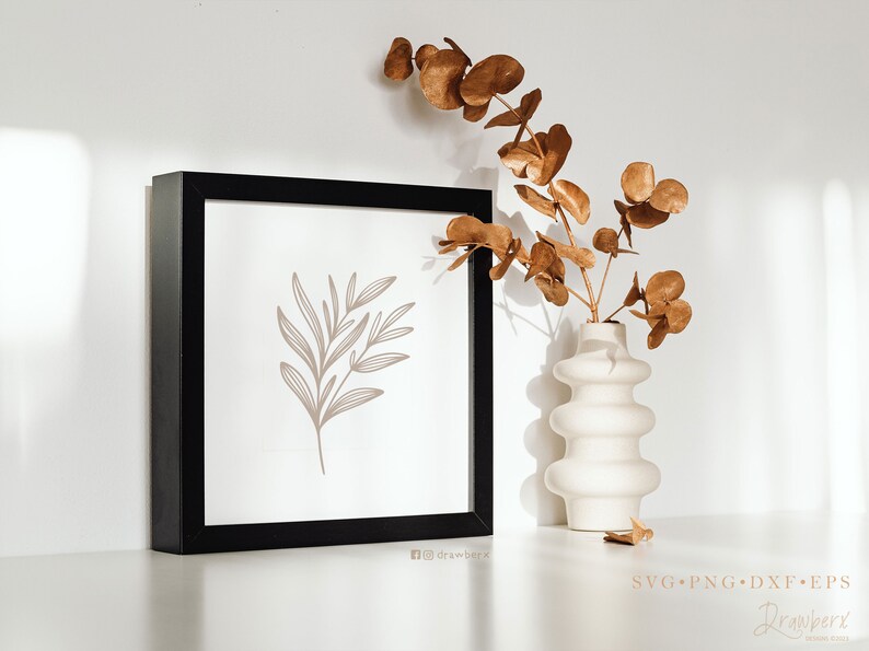 May include: A black framed print featuring a simple line drawing of a branch with leaves in a light brown colour. The print is sitting on a white surface next to a white vase with dried brown eucalyptus branches.