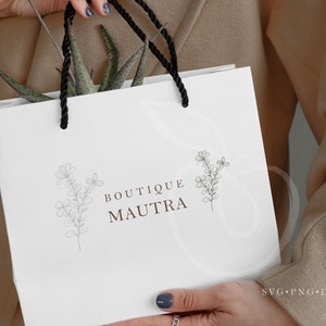 May include: White paper shopping bag with black handles and a logo that says "Boutique Maufra" with a floral design on either side of the text.