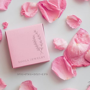 May include: A pink box with the text "Xoola Jewelry" and a simple line drawing of a sprig of leaves. The text "SVG.PNG.DXF.EPS" is printed below the company name. The box is on a white surface with pink rose petals scattered around it.