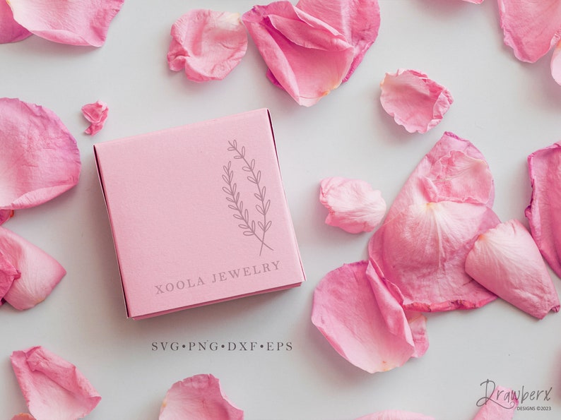 May include: A pink box with the text "Xoola Jewelry" and a simple line drawing of a sprig of leaves. The text "SVG.PNG.DXF.EPS" is printed below the company name. The box is on a white surface with pink rose petals scattered around it.