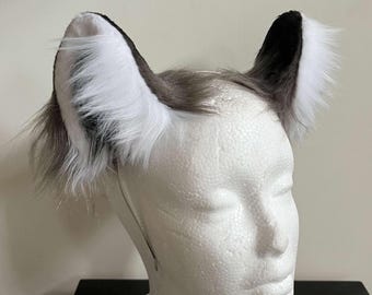Può includere: Un cerchietto con due orecchie da animale dritte realizzate in pelliccia sintetica a pelo lungo. Le orecchie presentano un motivo a gradiente di colore, che passa dal grigio scuro alla base e all'interno al bianco brillante sui bordi esterni.