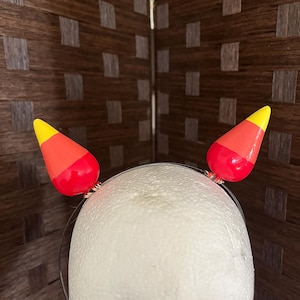 May include: A Halloween-themed headband featuring two candy corn decorations. Each candy corn is painted with yellow, orange, and red colors. The headband is made of a thin, silver-colored metal.