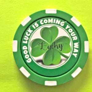 Op de afbeelding: Een groene en witte pokerchip met de tekst "Good Luck Is Coming Your Way" rond de rand. Het midden toont een groene klaver met vier bladeren met het woord "Lucky" in cursief.