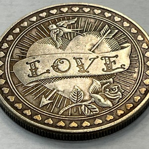 May include: Silver-toned round coin with a heart pierced by an arrow and a banner reading "LOVE". The heart is surrounded by a ring of small hearts.