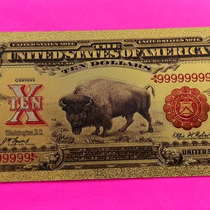 May include: A gold-colored replica of a $10 United States bill featuring a buffalo, a portrait of a man, and the text "UNITED STATES OF AMERICA" and "TEN DOLLARS".