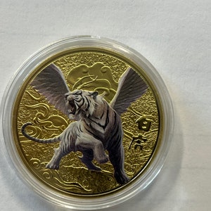 May include: A gold-colored coin with a white tiger with wings on a gold background. The coin has Chinese characters on it.