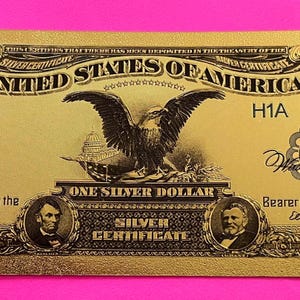 May include: A gold-colored replica of a one dollar bill featuring an eagle, the words "United States of America", and the text "One Silver Dollar Silver Certificate".
