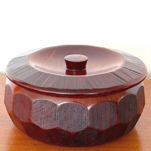 May include: A handcrafted wooden bowl with a lid. The bowl is a rich reddish-brown color and features a geometric design on the sides. The lid has a small, rounded knob and a textured surface. This decorative bowl is ideal for storing small items.