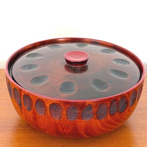 May include: A round, wooden bowl with a matching lid. The bowl and lid are stained a reddish-brown color with a dark brown pattern. The lid has a small, red knob in the center. The bowl is sitting on a wooden surface.