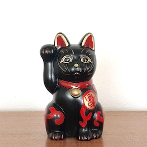 May include: A black Maneki-neko figurine with a raised paw, a traditional Japanese lucky cat. The cat has red accents on its ears, collar, and decorative markings. A gold-colored bell hangs from its collar. The figurine is on a wooden surface.