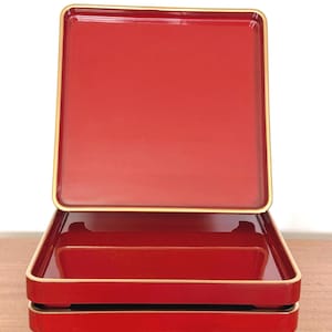May include: A set of two square, glossy red lacquer trays with rounded corners and thin gold-colored trim along the edges. The trays are stacked, with the top tray positioned vertically and the bottom tray resting horizontally on four small feet.