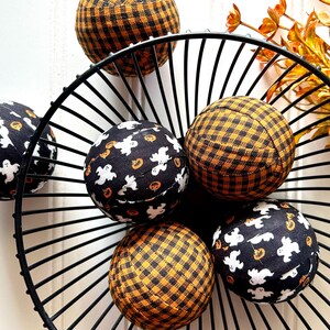 May include: Six Halloween-themed fabric balls in a black metal wire basket. Three balls feature a black and orange buffalo plaid pattern, and three have a black background with white ghosts and orange pumpkins. These decorative balls are perfect for autumn and Halloween decor.