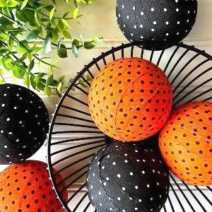 May include: A decorative arrangement featuring black and orange polka-dot spheres. Some spheres are nestled in a black wire basket, while others are placed around it. Green foliage adds a natural touch to the display.