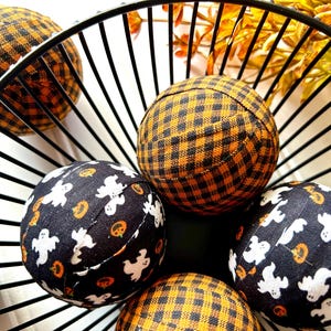 May include: A black wire basket filled with fabric balls. The balls are covered in black and orange plaid fabric and black fabric with white ghost and pumpkin designs.