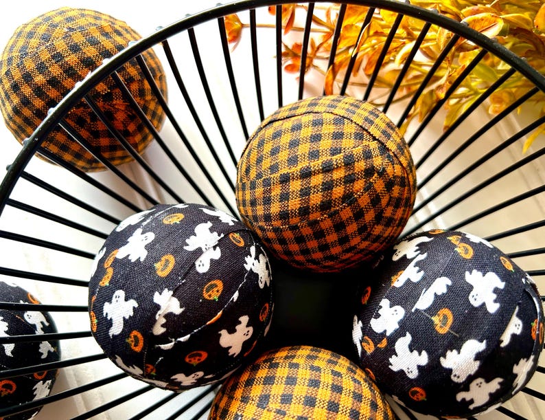 May include: A black wire basket filled with fabric balls. The balls are covered in black and orange plaid fabric and black fabric with white ghost and pumpkin designs.