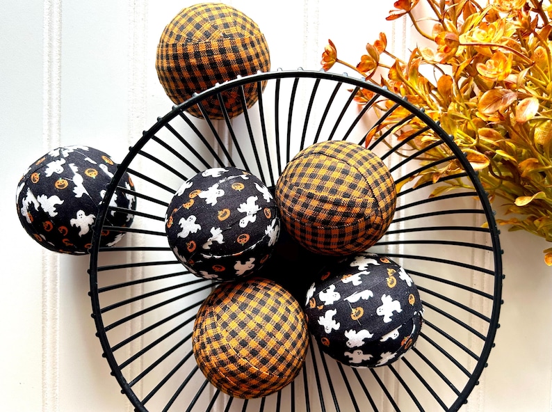 May include: Six Halloween-themed fabric balls in a black metal wire basket. Three balls feature a black and orange buffalo plaid pattern, and three have a black background with white ghosts and orange pumpkins. These decorative balls are perfect for autumn and Halloween decor.