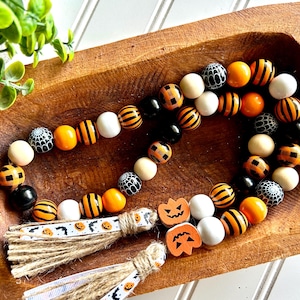 May include: A Halloween-themed wooden bead garland with orange, black, and white beads. The garland features pumpkin and spiderweb designs, with jute tassels and ribbon accents. The beads are arranged in a decorative wooden bowl.