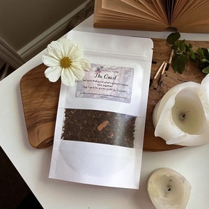 Puede incluir: Una bolsa de té blanca con la etiqueta "The Quest" con hojas de té y cáscara de naranja visibles. La bolsa está sobre una tabla de madera con una margarita blanca, una vela blanca parcialmente quemada y un libro abierto.