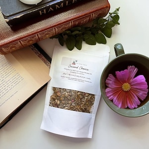 Puede incluir: Naturaleza muerta con una bolsa blanca de té "Second Chance", una taza de té con una flor rosa y dos libros antiguos. Una vela blanca está encima de los libros. La bolsa de té tiene el texto "Following your heart isn't always the easy, but sometimes it's worth it."