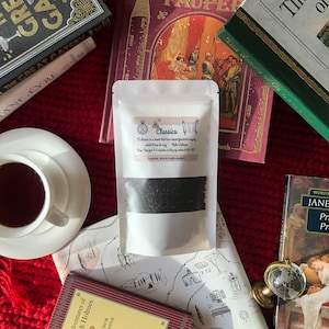 Può includere: Un sacchetto bianco di tè sfuso con l'etichetta "Classics" con una finestra trasparente, circondato da libri e una tazza da tè. L'etichetta include una citazione e le istruzioni per l'infusione. La scena è ambientata su una superficie a maglia rossa, con una mappa e un piccolo globo.